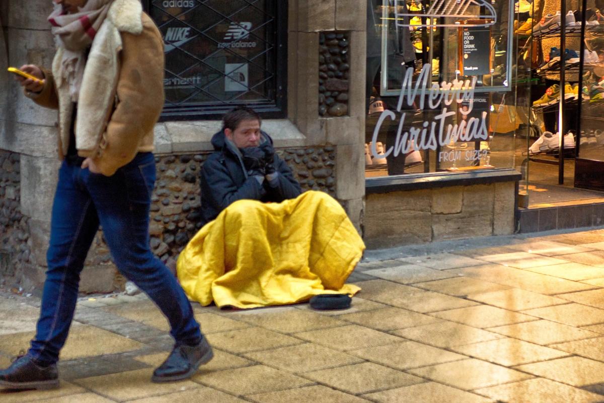 A homeless man sits on the street outside a shop with a Merry Christmas sign in the window