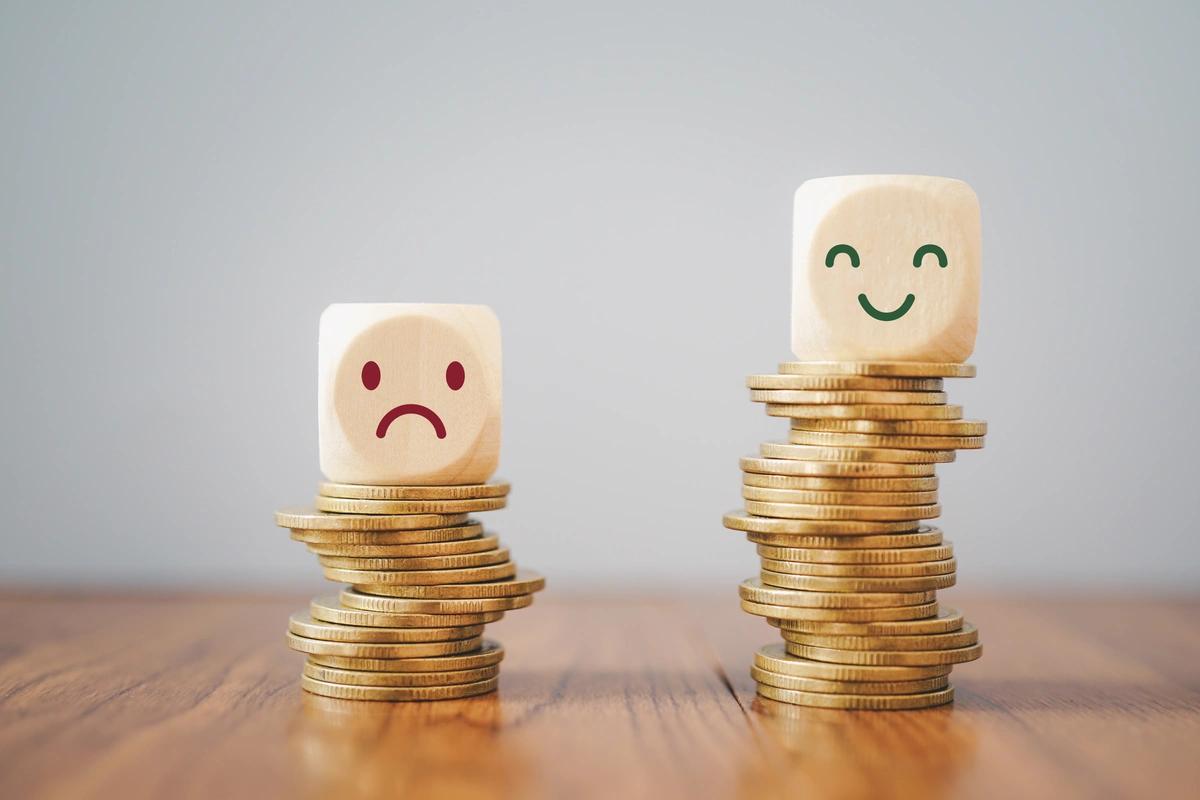 Two cubes each sitting on a stack of coins. The cube sitting on the higher stack has a smiley face and the cube sitting on the smaller stack has a sad face.