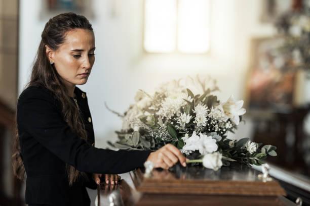 Image of a coffin with a floral display on top and a sad looking woman standing over it place a single flower on top