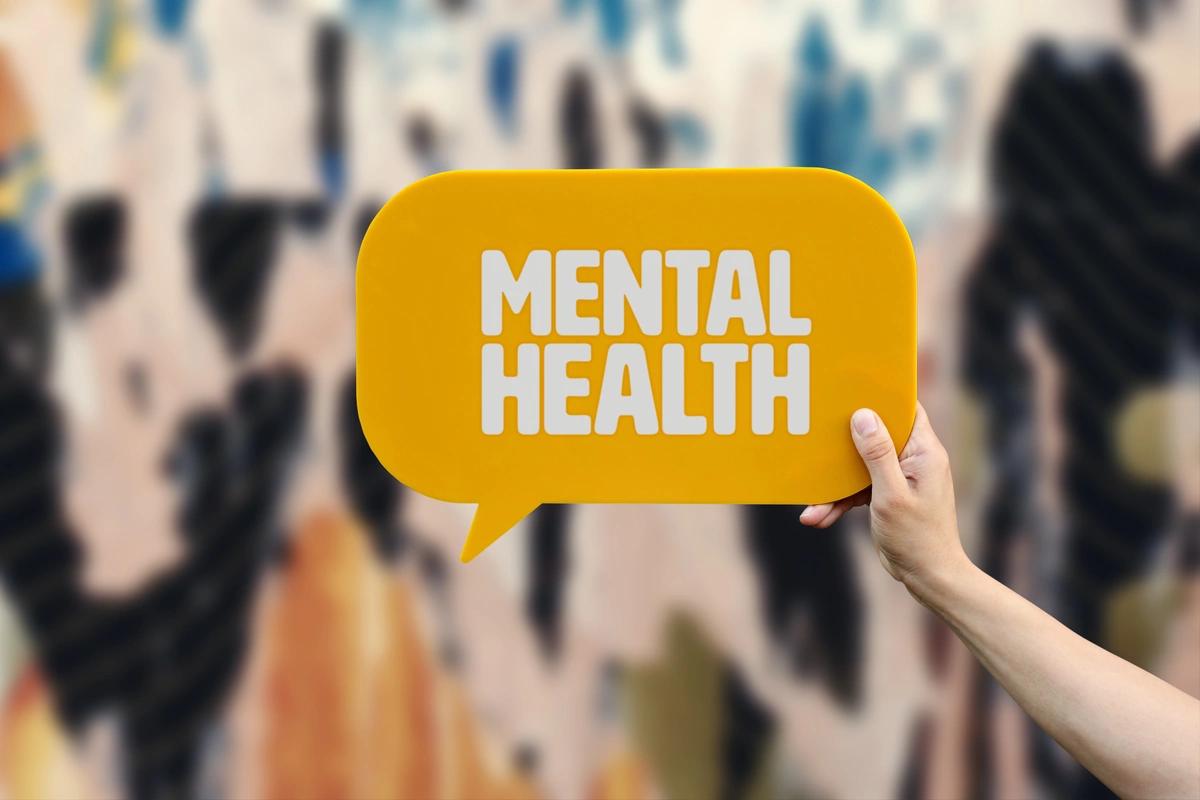 A hand holds up a yellow speech bubble sign with the words 'mental health' written on it