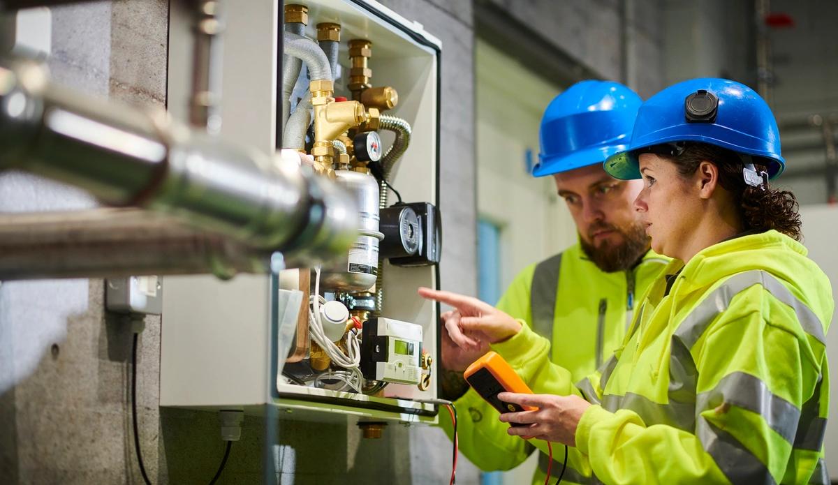 Two engineers in blue hard hats and yellow high-vis jackets examine an energy meter