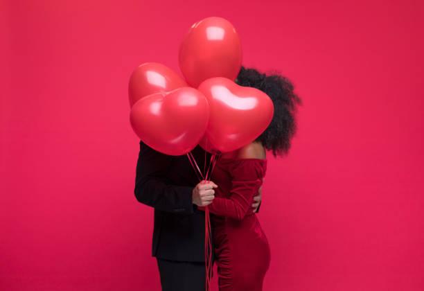 Image of a couple kissing behind a bunch of red heart balloons. 60% of people spending less on Valentine's Day due to cost of living crisis