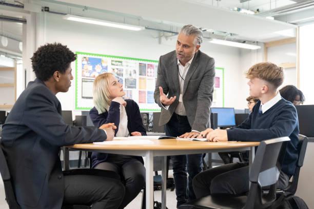 Teenage school children sat around a desk talking with their teacher