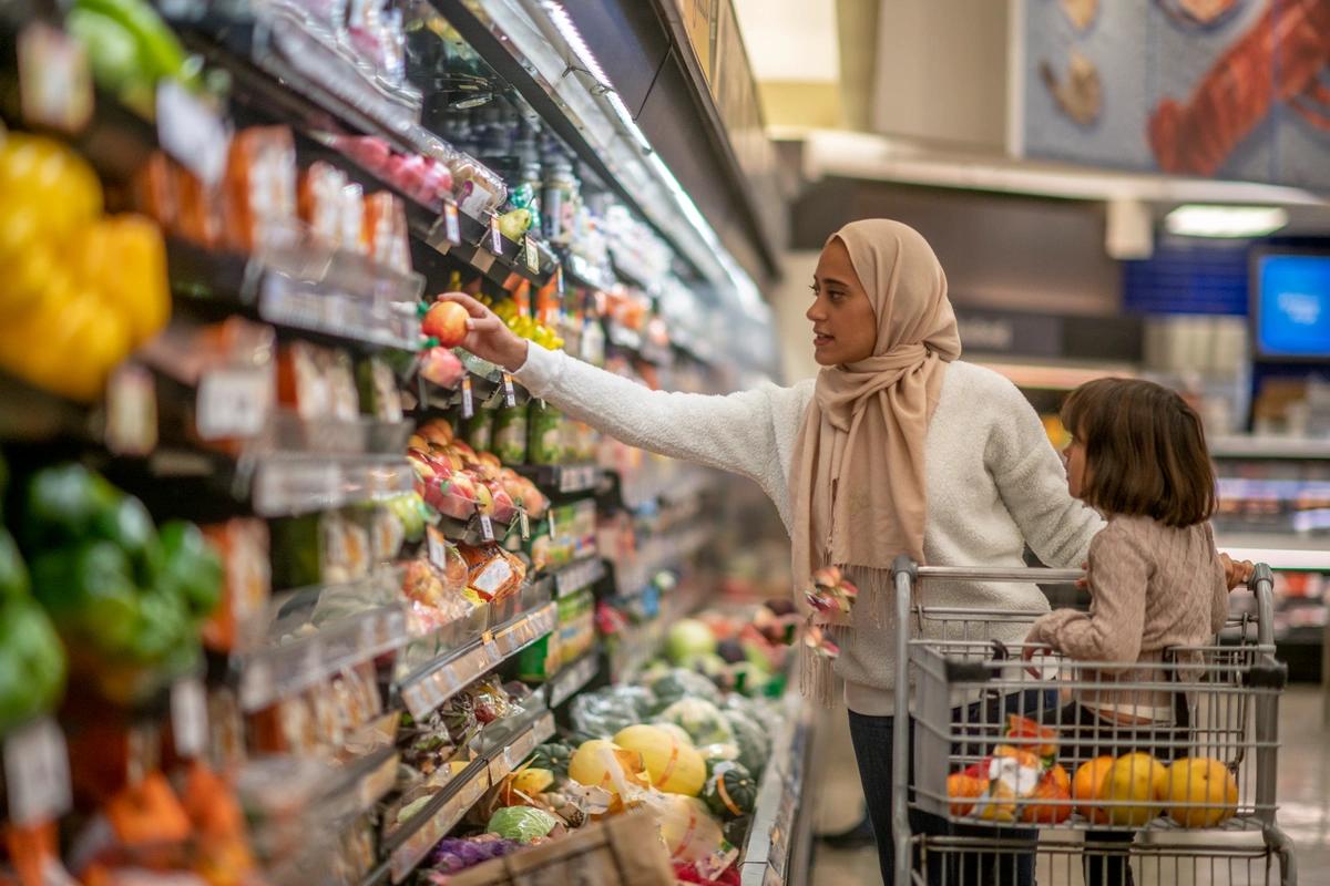 A woman selects food items from a supermarket fridge while her young daughter looks on from the trolley