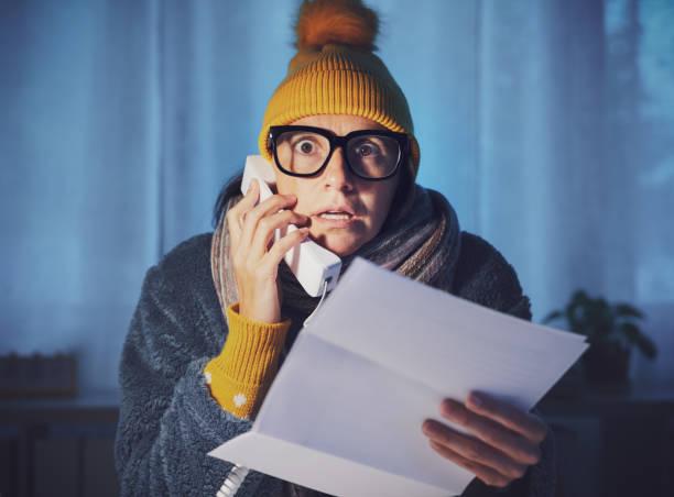 Image of a woman with a woolly hat on looking cold on the phone to her energy provider with a bill in her hard. Household support fund - how it could help with energy bills