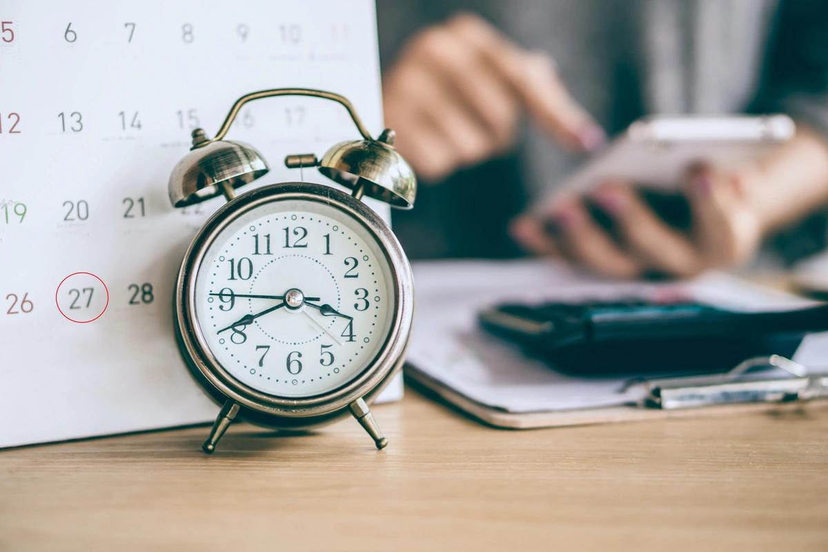 Clock on desk next to calendar and person calculating on phone