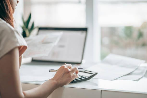 Image of a woman with a laptop open and a budget spreadsheet in front of her, looking at bills and using a calculator