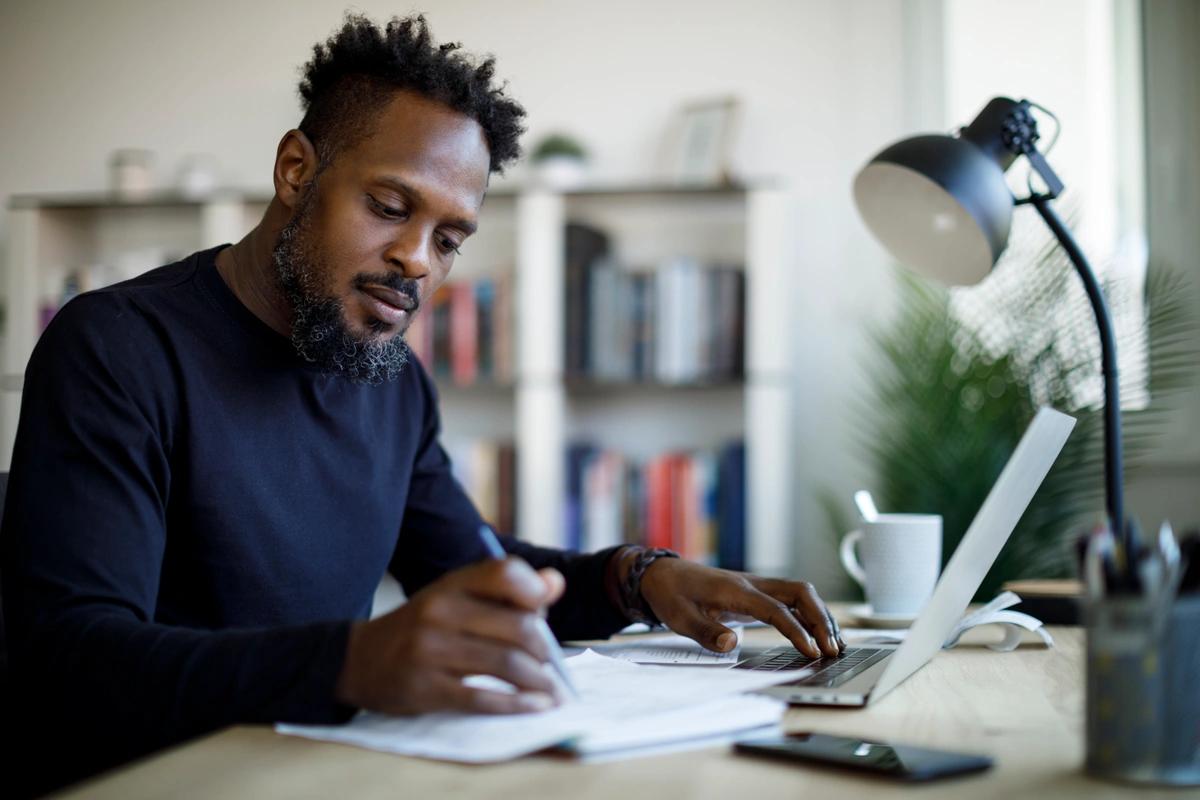 Man sitting at a desk in front of a laptop jotting things down on a pad of paper