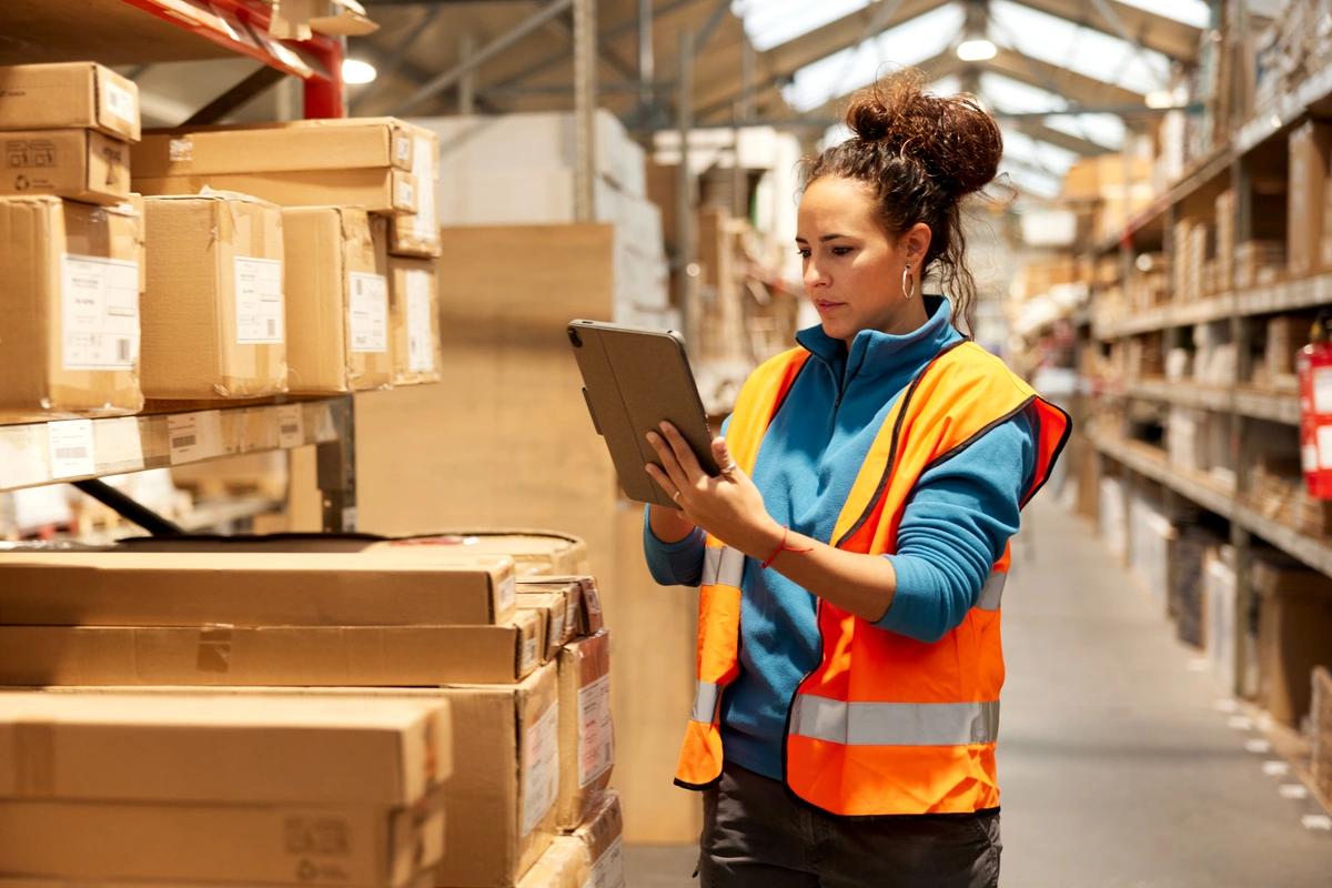A woman in a high-vis vest stands in a warehouse writing on a clipboard