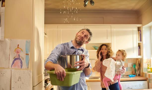 Image of a man holding a bucket under a leaking ceiling with a woman in the background holding a baby while on the phone. Home insurance prices rise by 32%. How to reduce your home insurance premium