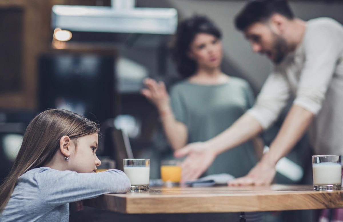 A little girl sits looking sad at the breakfast table while her parents argue