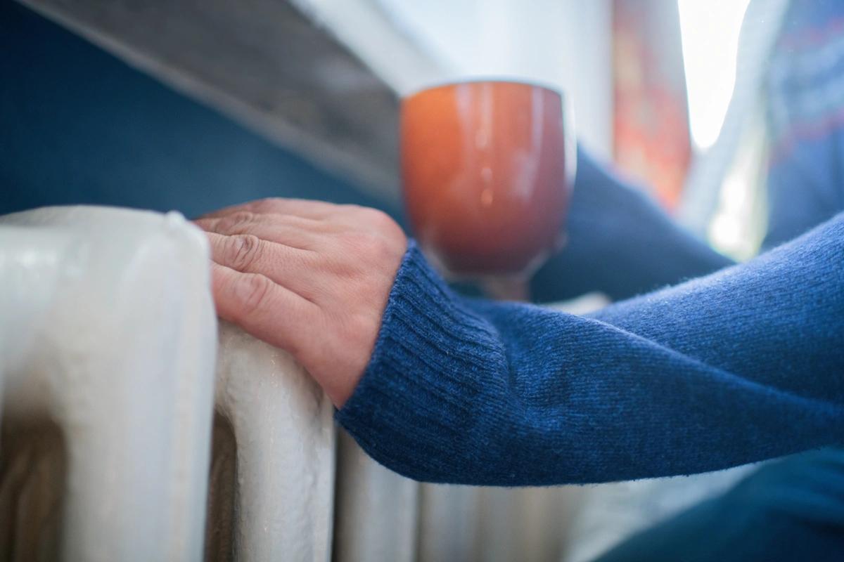 A person tries to get warm by a radiator with a cup of tea