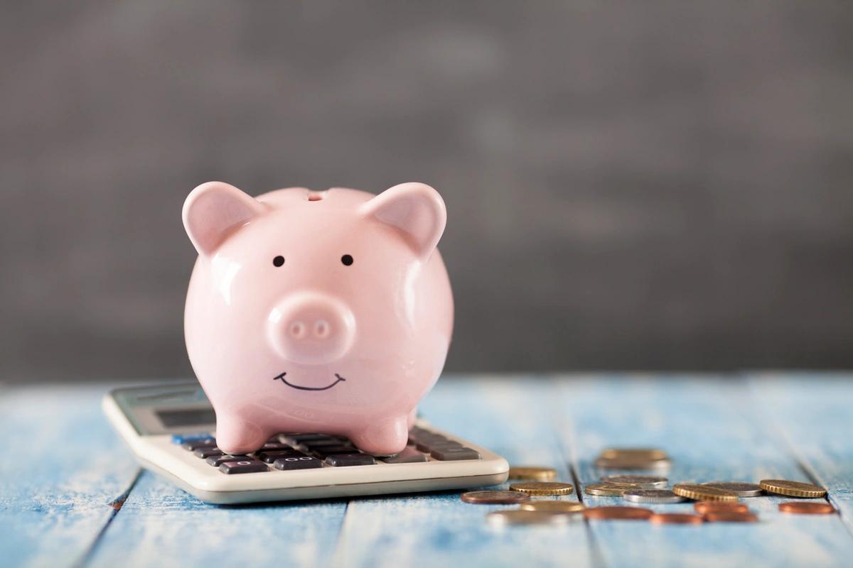 A smiling piggy bank sits on top of a calculator with cash beside it