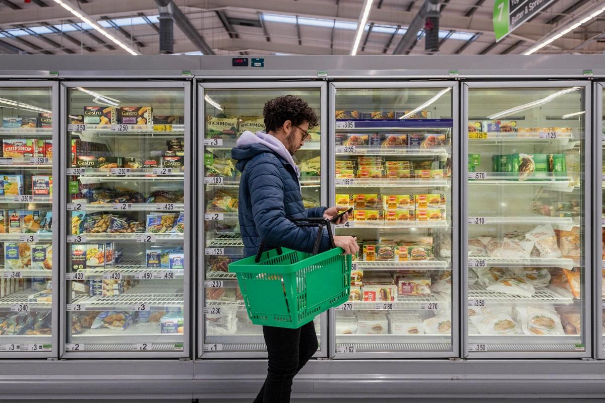 A man with a basket shopping in a supermarket stands looking at his phone in front of a row of freezers