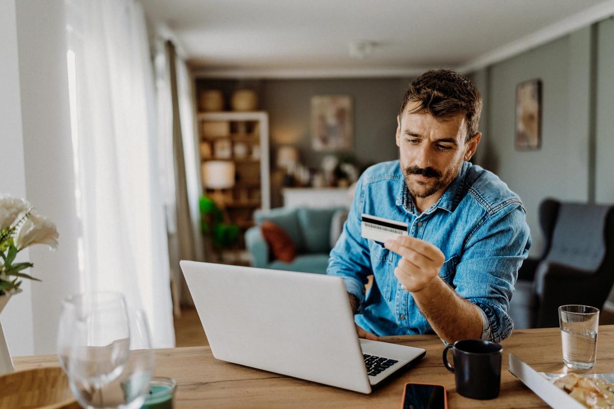 A man sits at his laptop with a credit card in his hand
