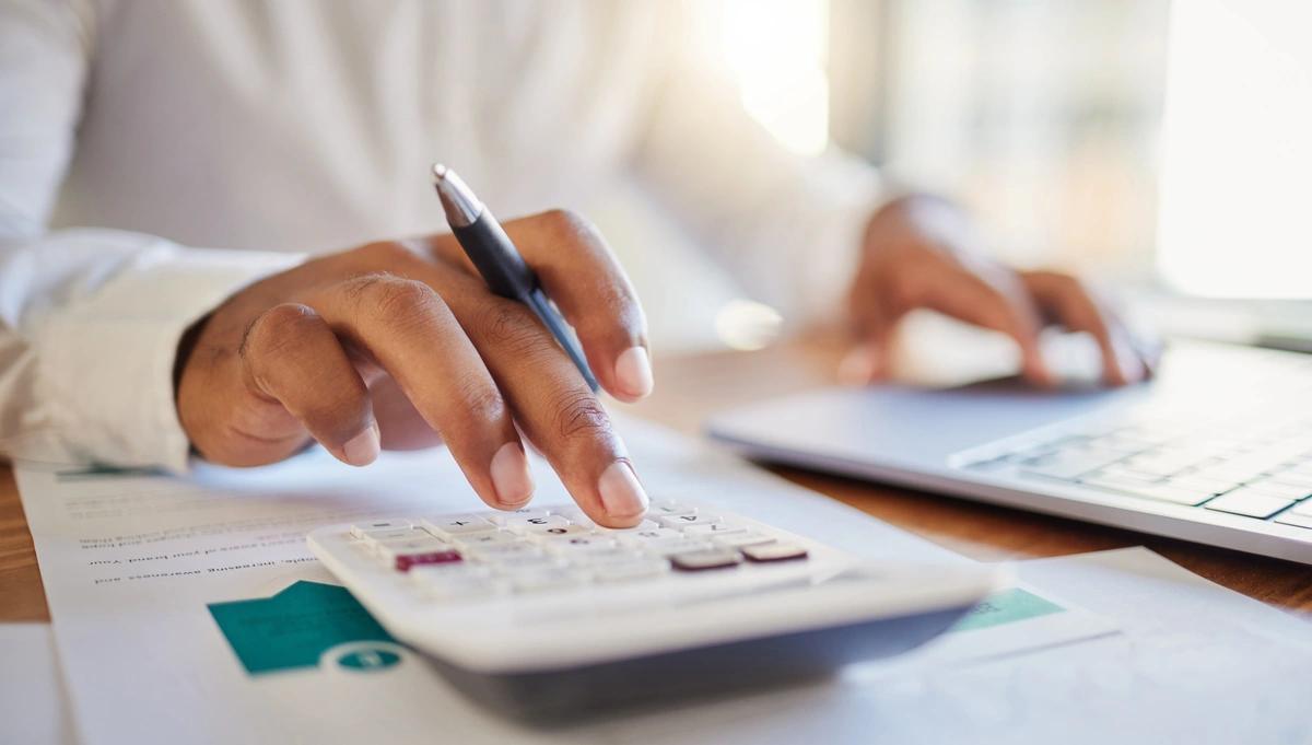 Close-up of a woman's hands as she works out her finances using a calculator and laptop