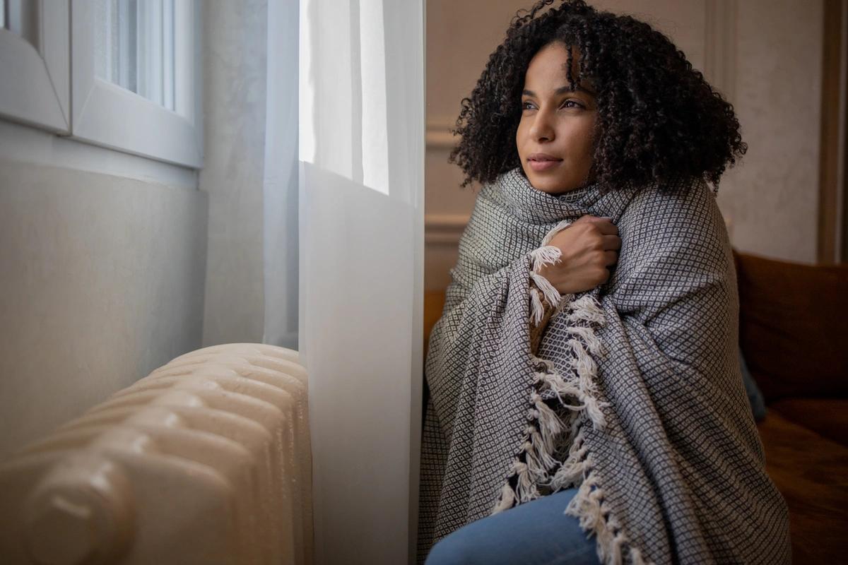 A woman wrapped in a blanket and huddling next to a radiator