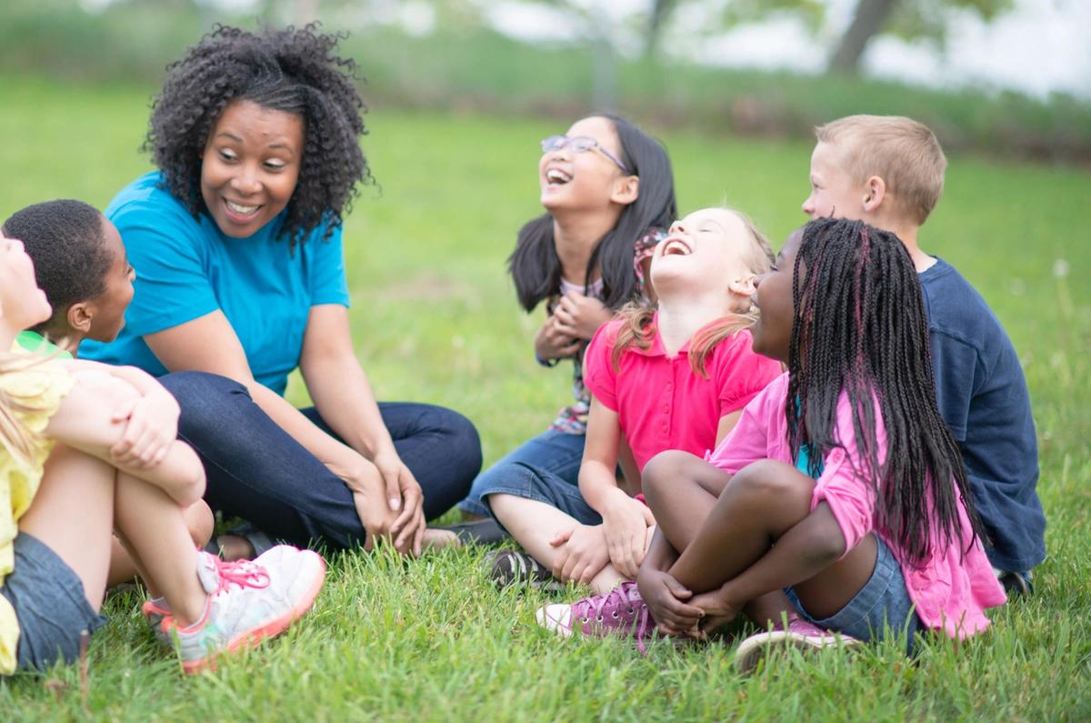 A group of laughing children sit in the grass with a childminder