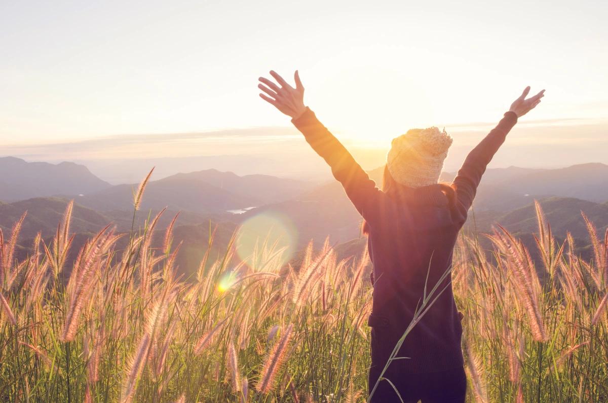 Shot of the back of a woman with her arms raised in triumph standing in a field at sunrise looking across a valley