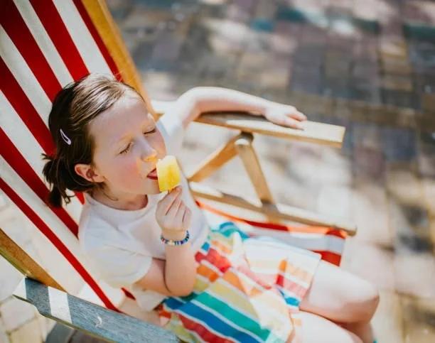 Picture of a little girl sat in a striped deckchair licking an ice lolly. 11 free ways to stay cool in the heatwave using things you already have at home. Free ways to stay cool and keep energy bills down