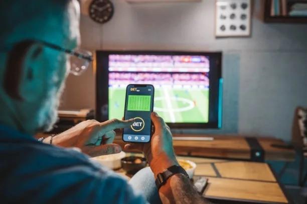 Image of a man sat in front on the TV with sport on - one his phone placing a bet. Gambling harm is being missed warns new report - the debt warning signs you shouldn’t ignore. A new report reveals millions of people could be experiencing gambling ha