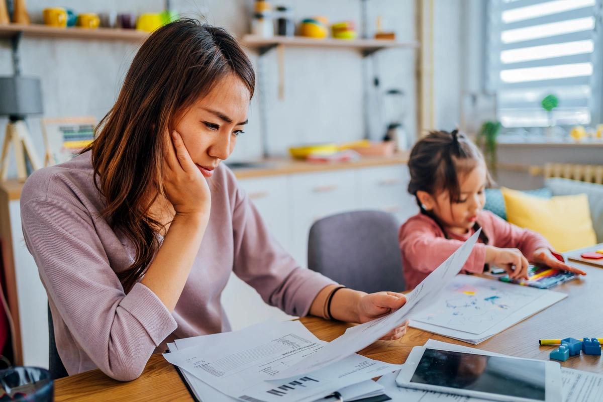 Mum going through bills at the kitchen table while her daughter draws a picture next to her