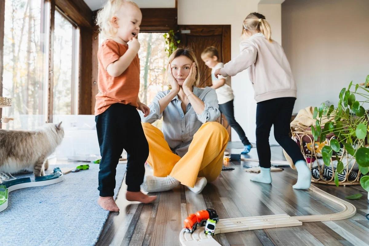 A stressed out mum sitting on the living room floor as her three kids run riot