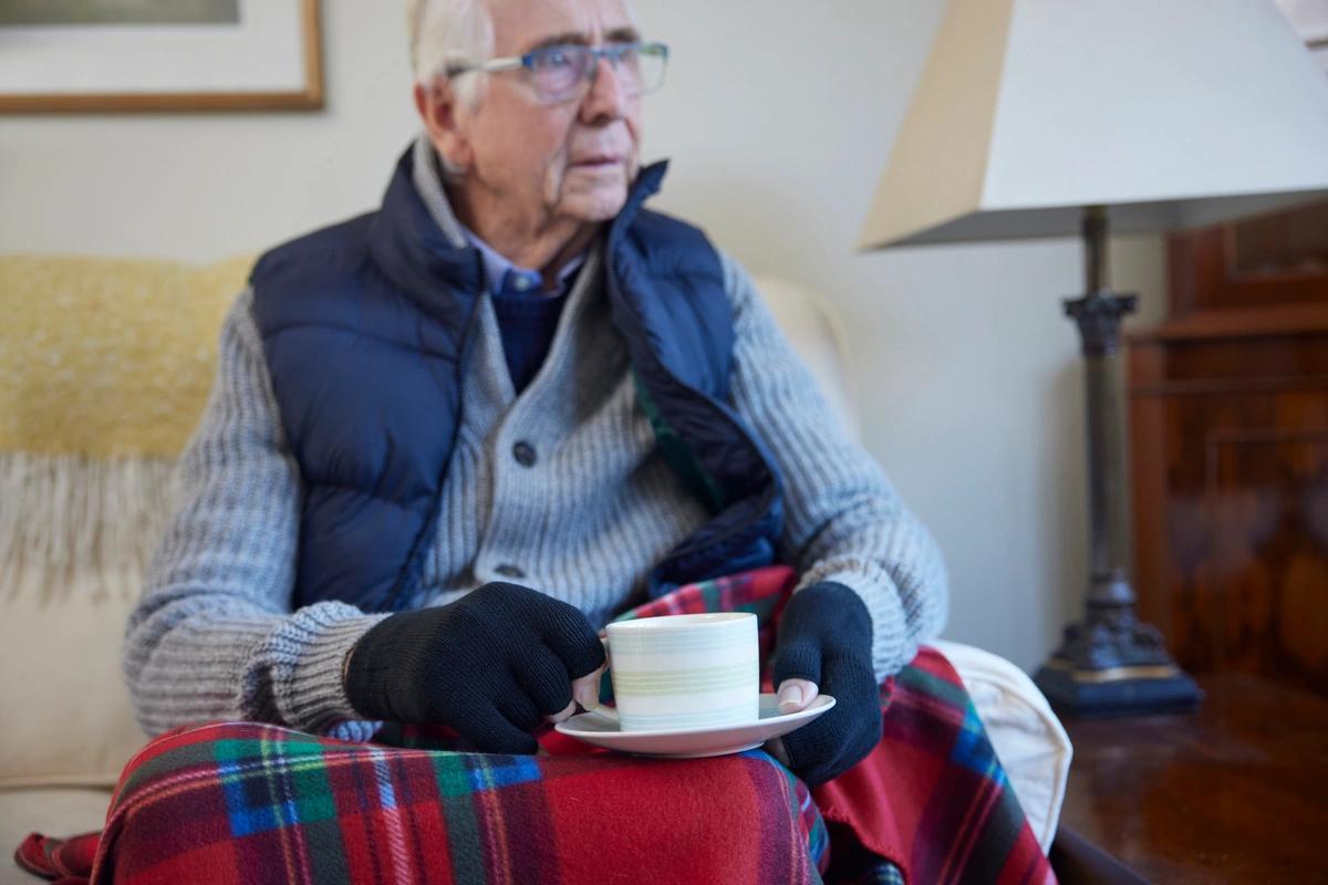 A old man wears extra clothes and drinks a hot drink to try and keep warm in his home