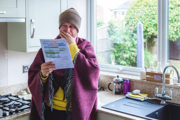 Image of a woman wrapped up in scarves, a hat and jumpers holding a bill and looking worried