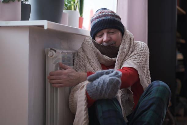 Image of a man wrapped up in layers of clothes with a scarf and hat on sat next to a radiator