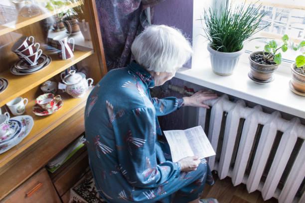 Image of a pensioner sitting next to a storage heater looking cold while reading a bill. Nearly 10 million households live in cold homes