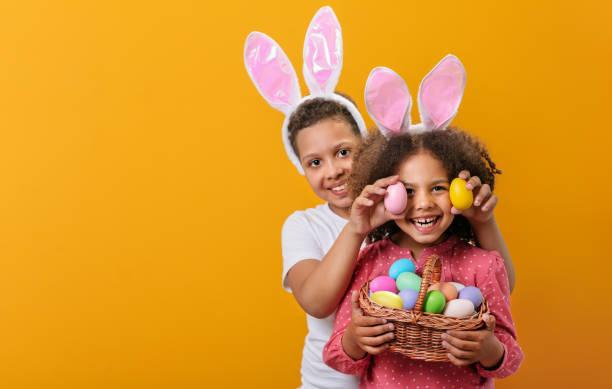 two children wearing bunny ears holding a basket of multi coloured Easter eggs