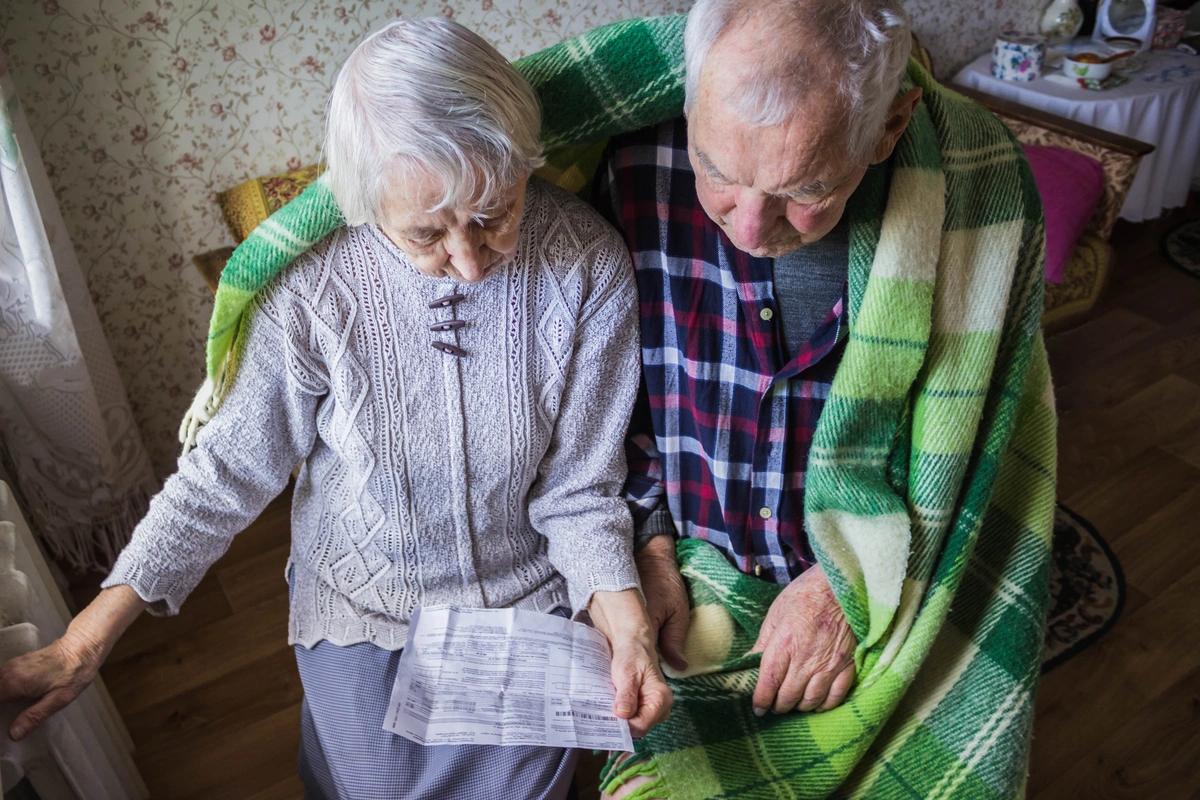 An elderly couple try to keep warm under a blanket and sitting next to a radiator while looking at their energy bill
