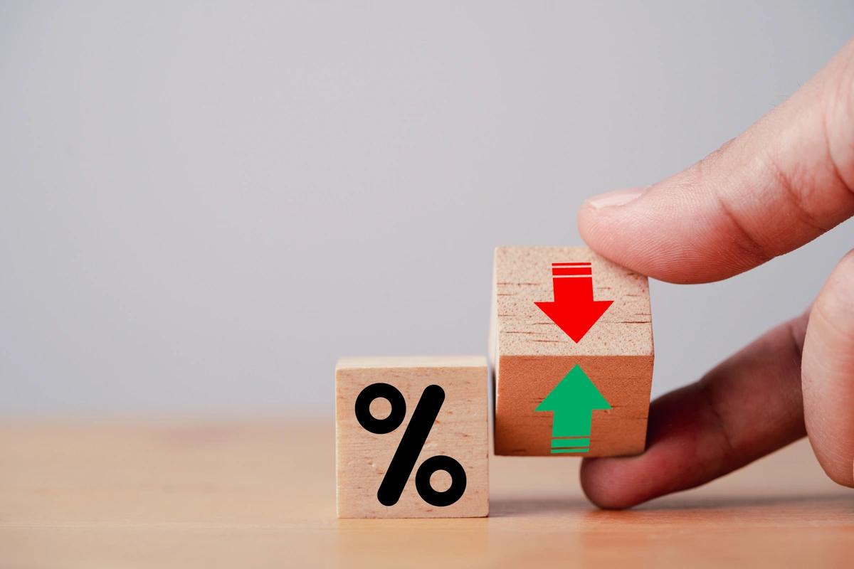 A hand tilting a wooden block with a red down arrow and a green up arrow next to a block with a percentage sign to represent the interest rate announcement