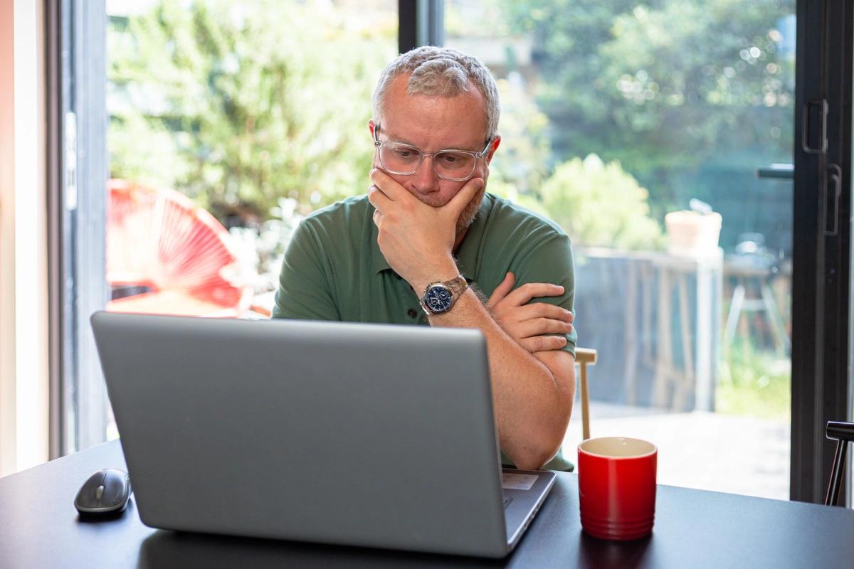 Serious-looking man sitting at a table in his home in front of a laptop