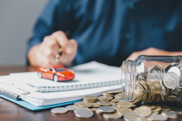 Image of a man writing in a note book with a toy car on top and a car full of coins falling over at the side to show ways to save on your car insurance premium
