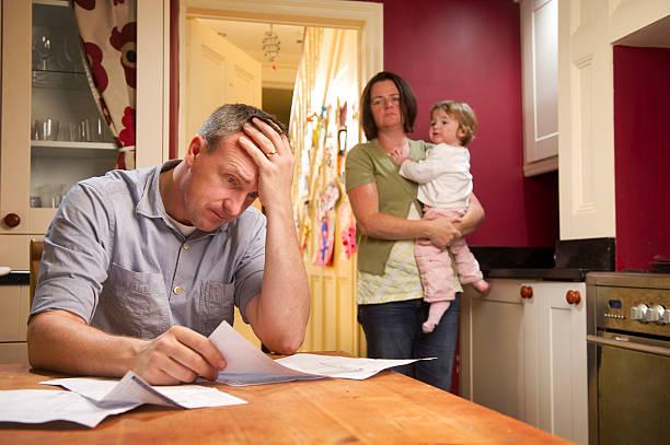 Image of a man with his head in his hands looking at bills with his wife stood behind him holding a baby. Five tips to getting out of debt