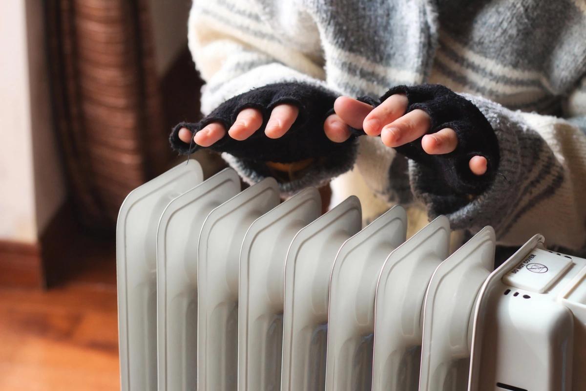 A person wearing a big jumper and fingerless mittens warms themselves over an electric heater
