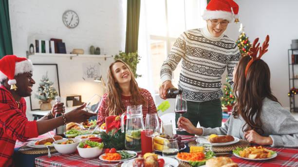 Image of three people sat around a table of Christmas food with party and santa hats on while a man serves red wine