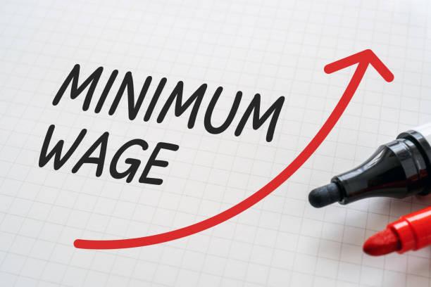 Image of 'minimum wage' written on a piece of paper in black with a red arrow pointing upwards as if to suggest it's going up