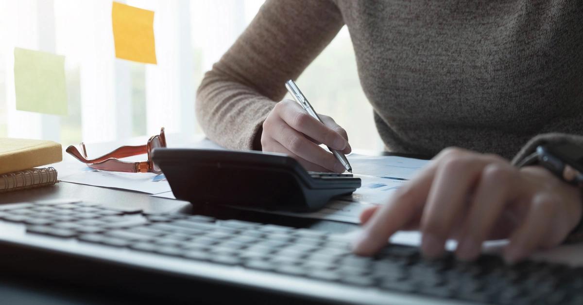 Close up of a woman using a calculator as she sits in front of a computer