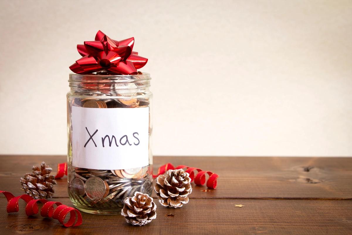A jar of coins with the word 'Xmas' written on it, a bow on the top and some pine cones scattered on the table next to it
