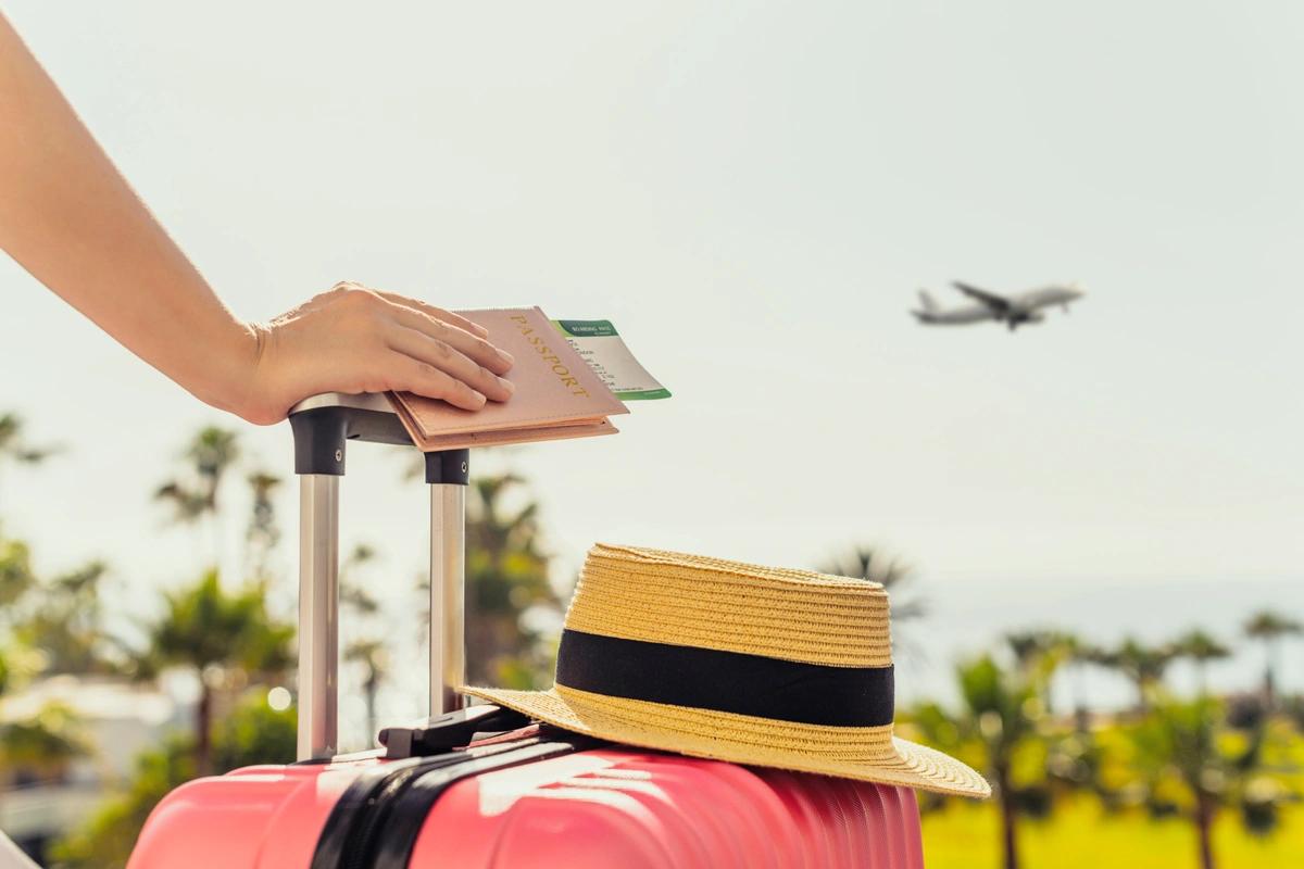 Close up of a woman's hand holding a boarding pass and leaning on a suitcase with palm trees in the background