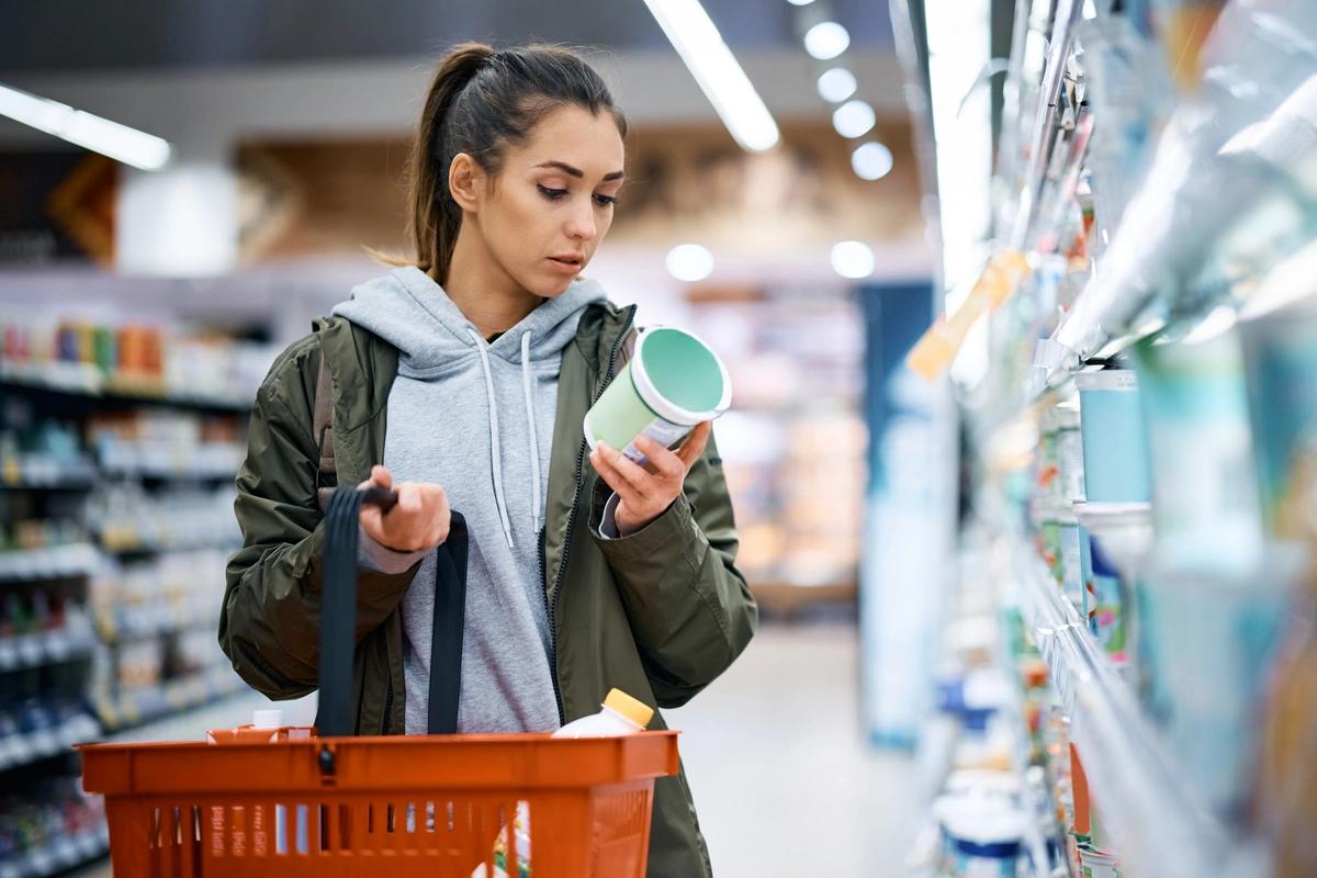 A woman with a shopping basket standing in a supermarket aisle reading the label of a food item she is holding in her hand