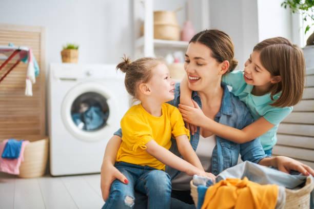 Image of a young mum sat on the floor sorting through the washing with a child hung over her shoulders and a toddler sat on her knee