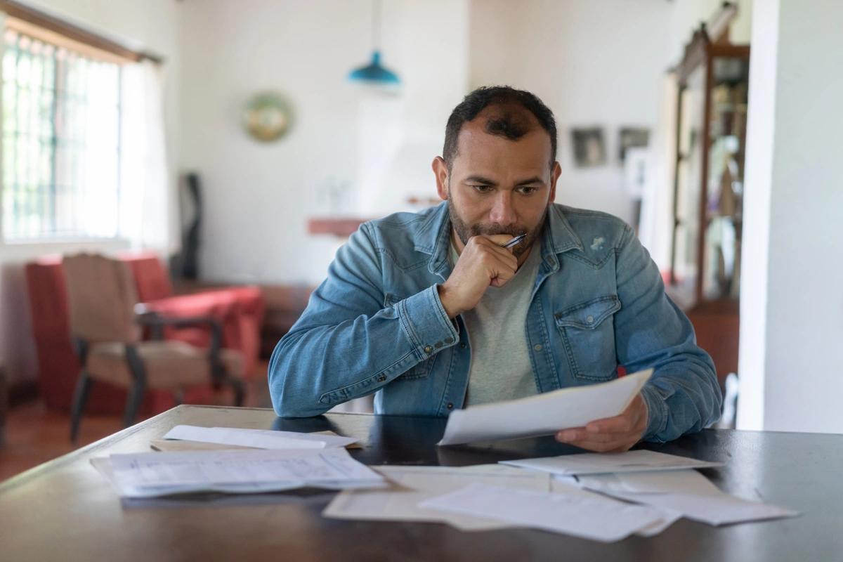 Man sitting at desk looking at bills with hand to his chin thinking